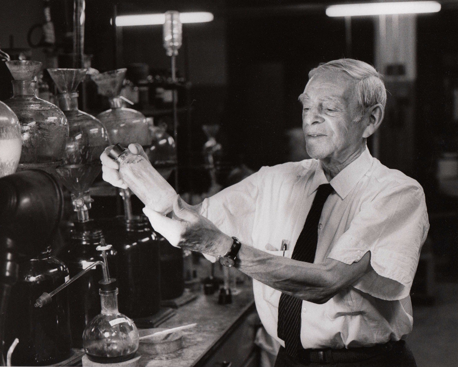 Black and white photograph of Casimir Funk holding a glass bottle with both hands with beakers in the background. 
