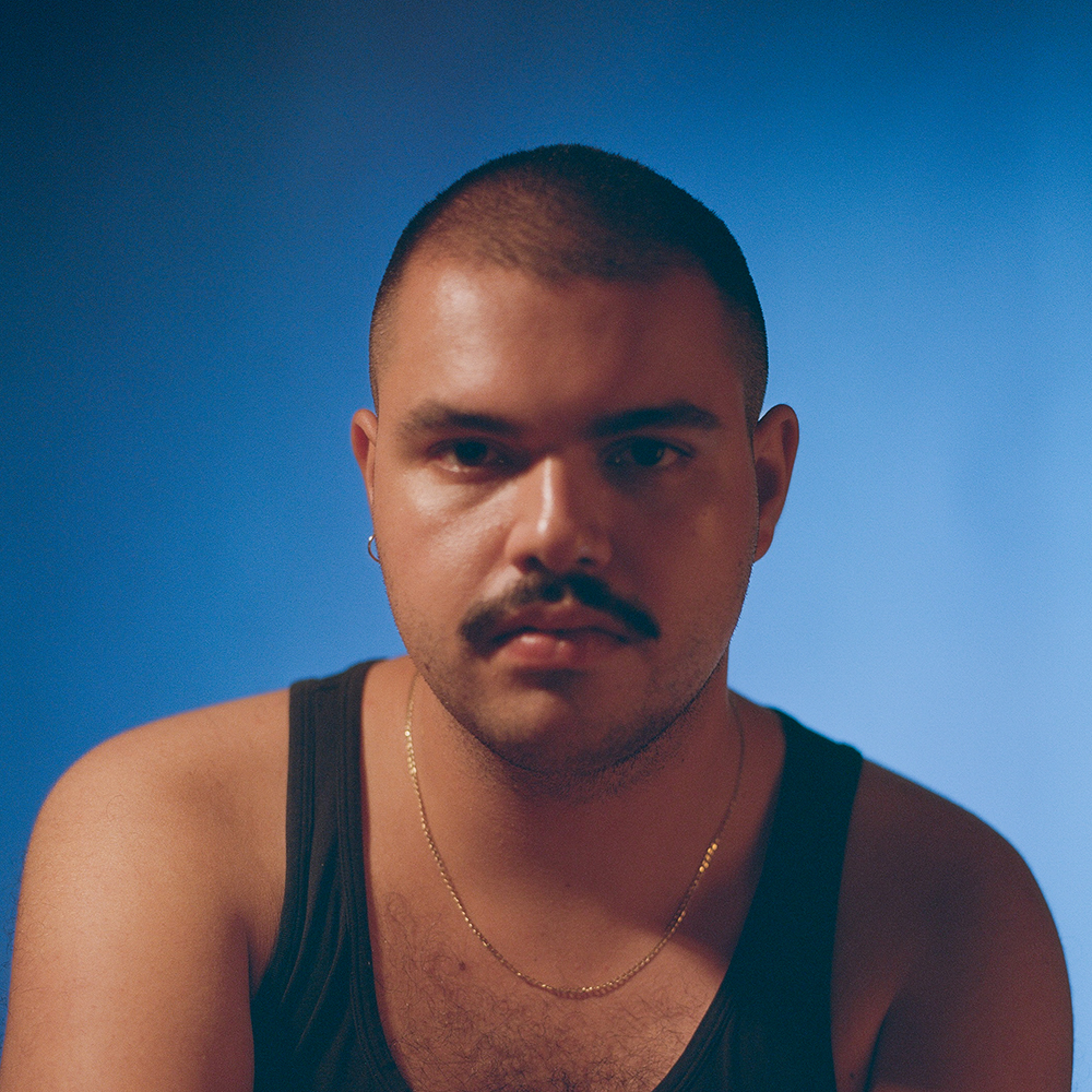 Photograph of a man with short black hair and a mustache looking directly to camera. He wears a chain necklace, hoop earring, and a black tank.