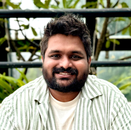 Photograph of an Indian man smiling with brown hair and beard. He's wearing a button up green and beige striped shirt with a beige t-shirt under and plants in the background.