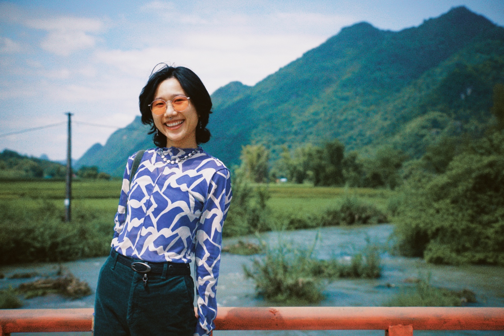 Photograph of a woman with short black hair and in a print shirt and round orange sunglasses. 