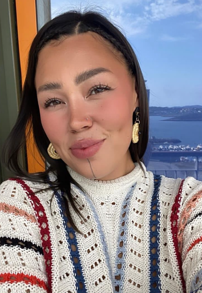 Photograph of Inuit woman wearing a striped sweater and gold earrings and straight black hair with two front pieces braided.