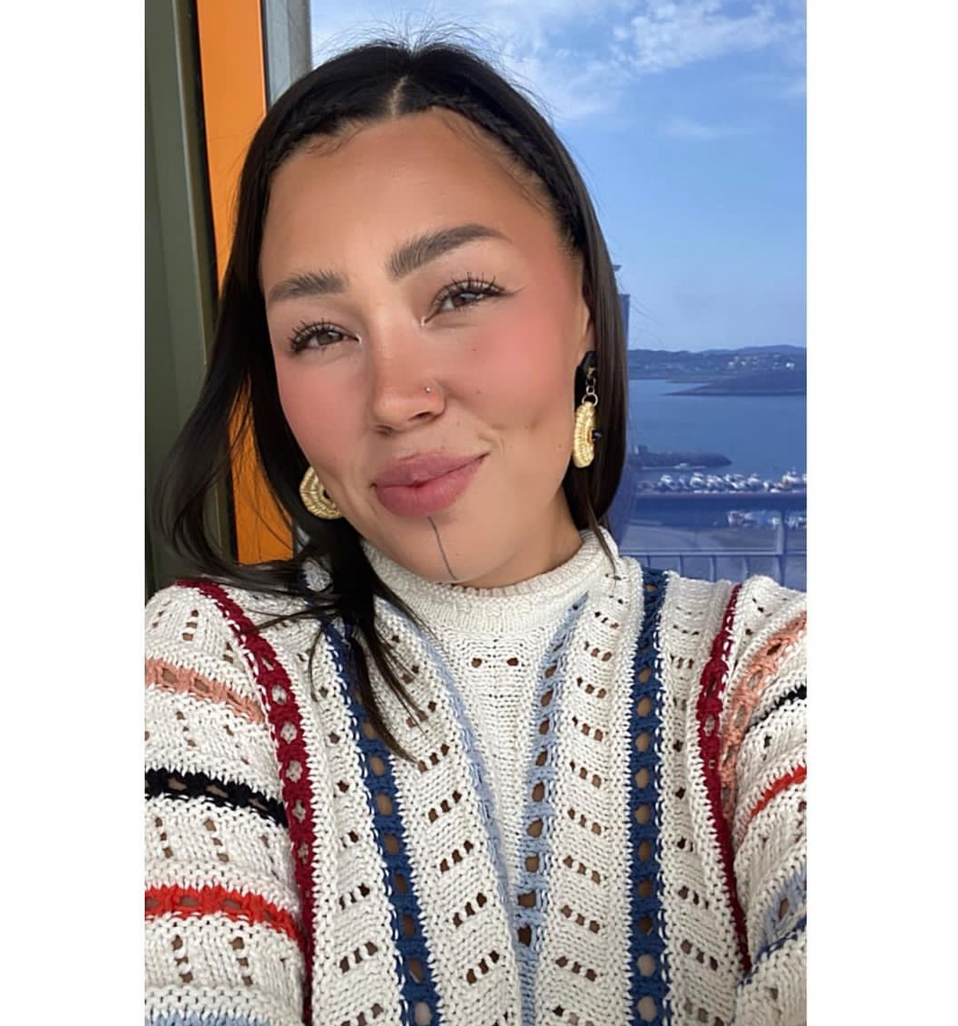 Photograph of Inuit woman wearing a striped sweater and gold earrings and straight black hair with two front pieces braided.