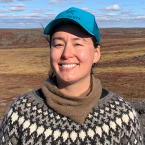 Photograph of Gayl  standing in front of a yellow and brown colored field outside with the sky showing above. She is smiling and has fair skin, dark brown eyes, and dark brown hair that is pulled back. She is wearing a blue ball cap, small hoop earrings, a light brown scarf and a crew neck sweater with a black and white diagonal pattern.