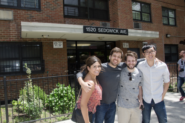 Photograph of 4 people hugging and smiling, standing in front of a brick building 