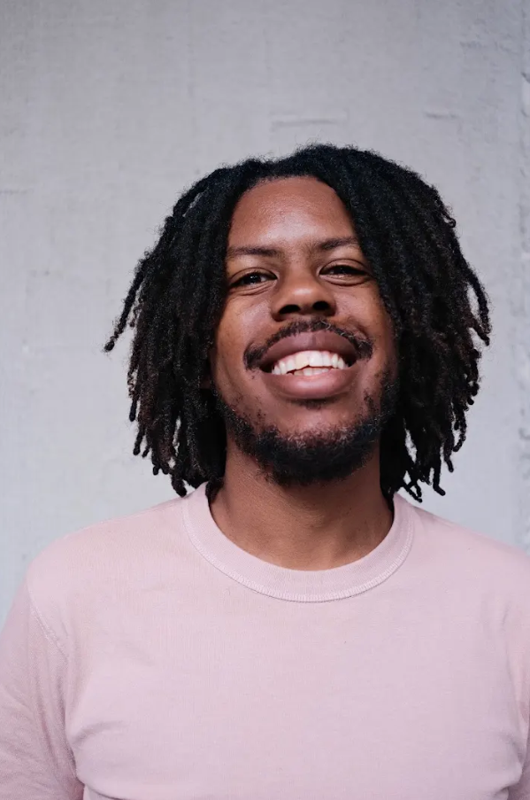 Photo of an African-American man with dreads and a pink shirt smiling into the camera