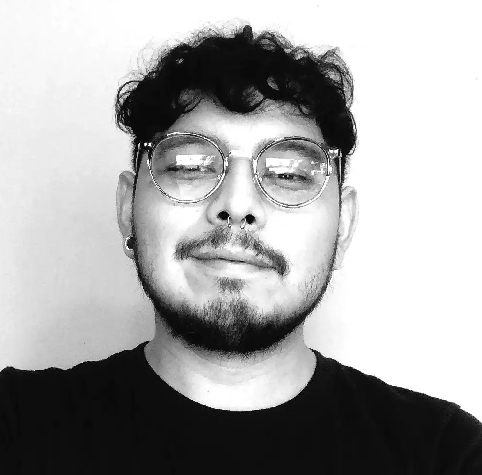 Black and white photograph of a hispanic man with short curly hair, wearing glasses and a black t-shirt smiling into the camera. 