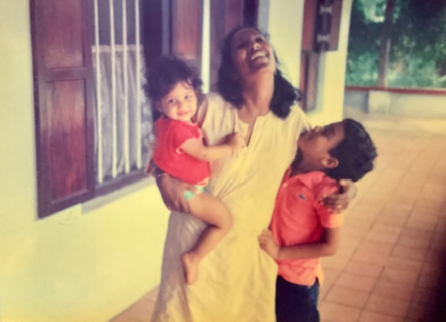 A Photograph of Meena laughing, holding one of her children in one arm, and wrapping her arm around her other child