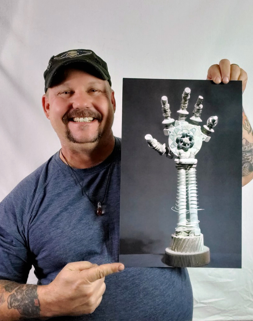 Photograph of a White man with facial hair smiling and pointing to a photo of metal sculpture of a hand. He's wearing a baseball cap and necklace and has tattoos on his forearm. 