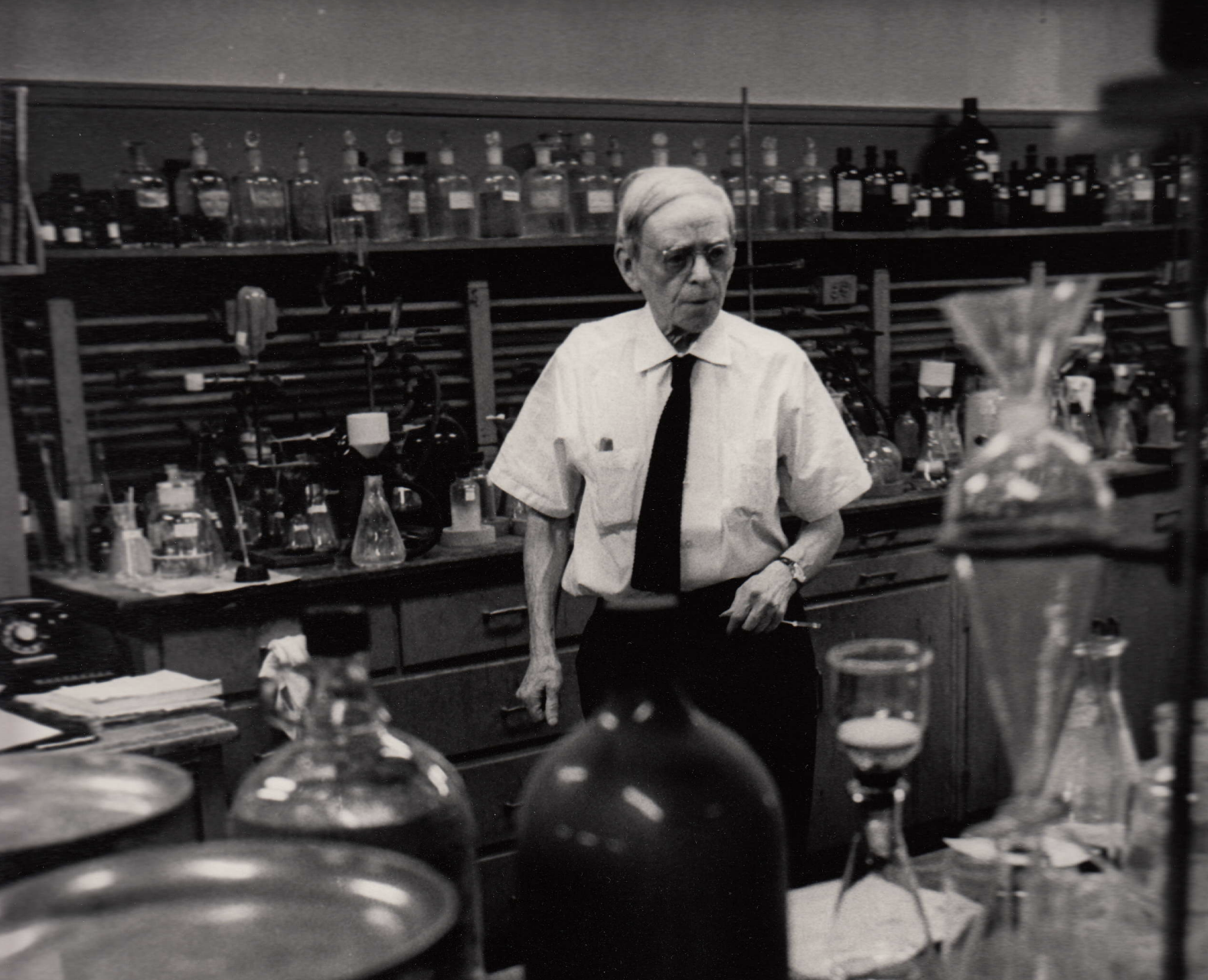 Black and white photograph of Casimir Funk  walking around a lab with many bottles and beakers in the fore- and background. 