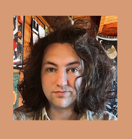 Color photo of Sonny, standing in front of a wall covered in art and photos. Sonny has voluminous, dark brown, wavy hair that is shoulder length. Sonny has fair skin, blue eyes, and has a faint grin. They are wearing a tan plaid collared shirt. 