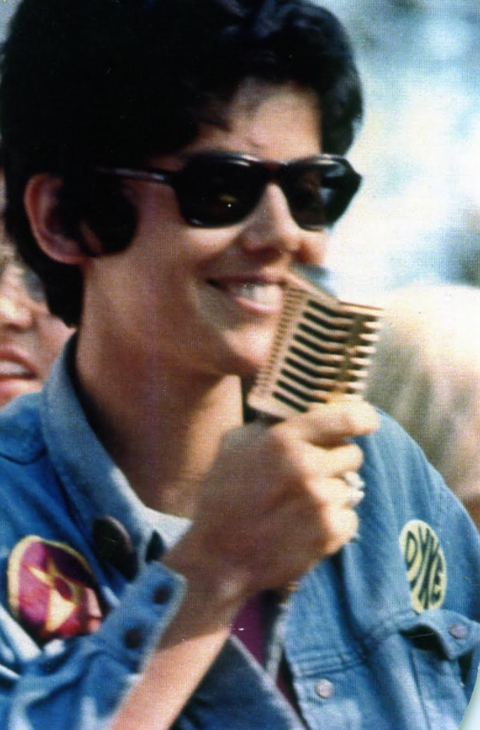 Photograph of Jeanne Córdova smiling, wearing sunglasses and holding a microphone.