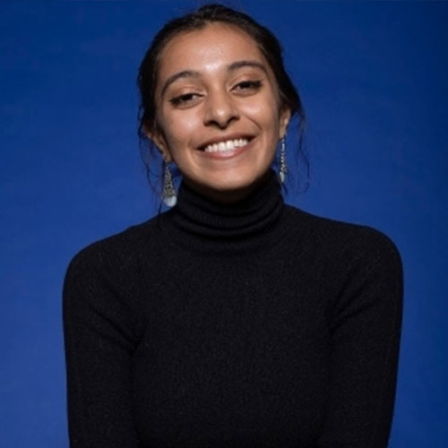 Color photograph of Tara smiling at the camera. She has black hair that is pulled back and dark eyes. She is wearing a black turtleneck and dangly silver earrings and is sitting in front of a dark blue backdrop.