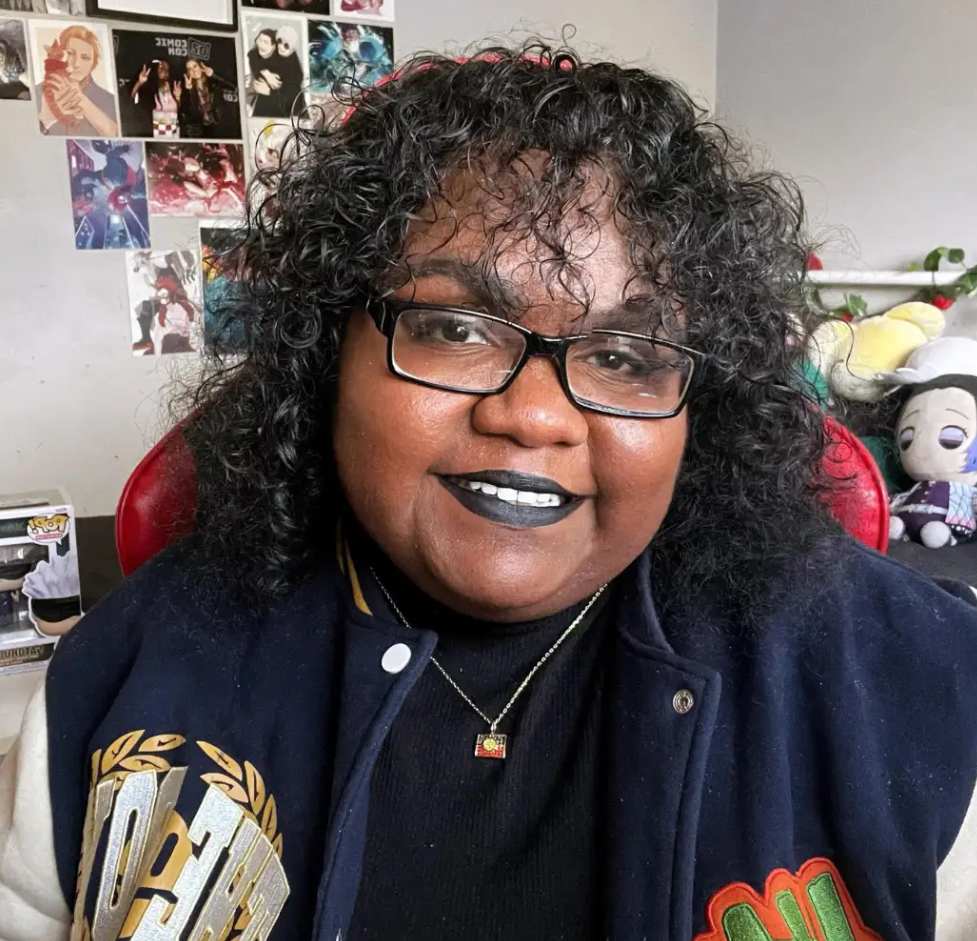 Photograph of Naarm woman wearing a navy blue and beige varsity jacket and a black shirt with short curly black hair sitting in a red chair. 