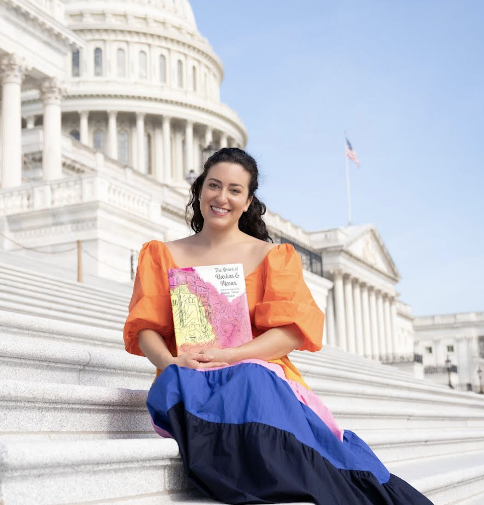 Photograph of a woman with long dark hair sitting on government building steps with the American flag waving in the background.