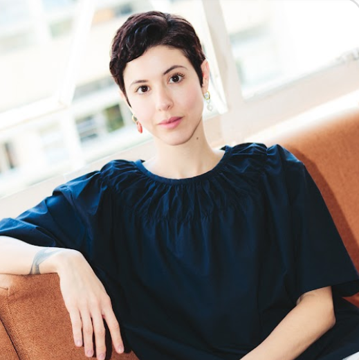 Photograph of white woman wearing a navy blue blouse and earrings, , sitting on a orange couch.