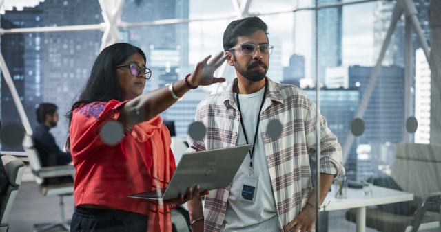 Woman with dark hair wearing glasses holding a laptop is showing a man with dark hear wearing glasses something on a clear board