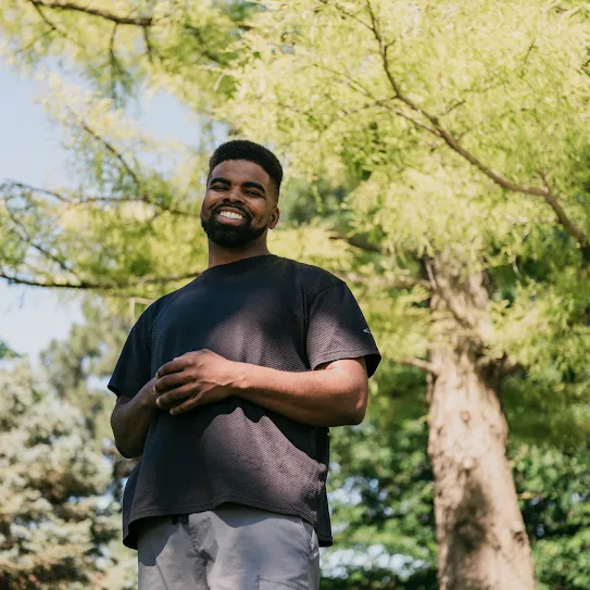 A recent Google Skilled Trades and Readiness Program graduate stands outside under the trees, smiling.