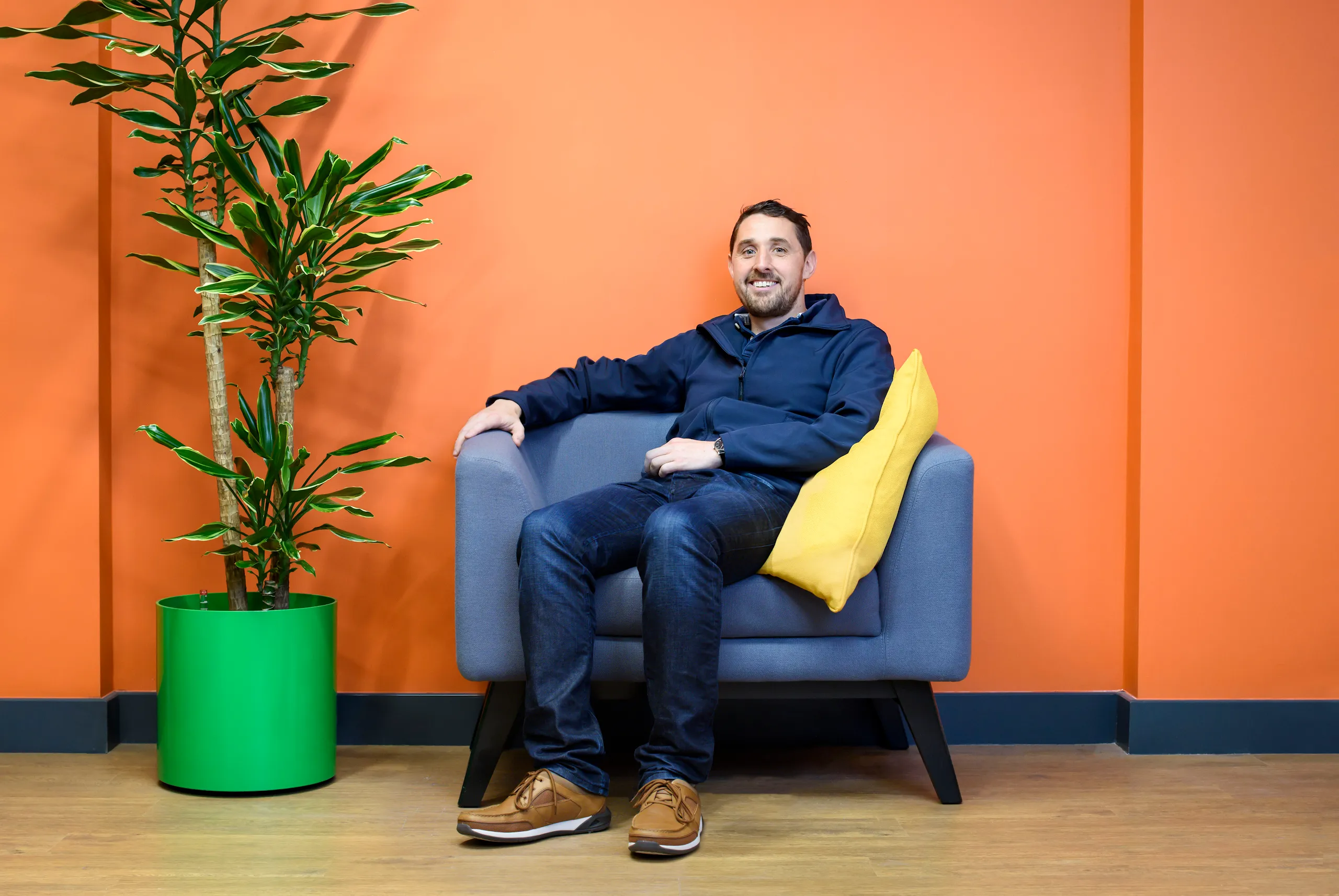 A photograph of a smiling man sitting in a chair next to a plant. 