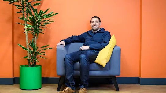 A photograph of a smiling man sitting in a chair next to a plant. 