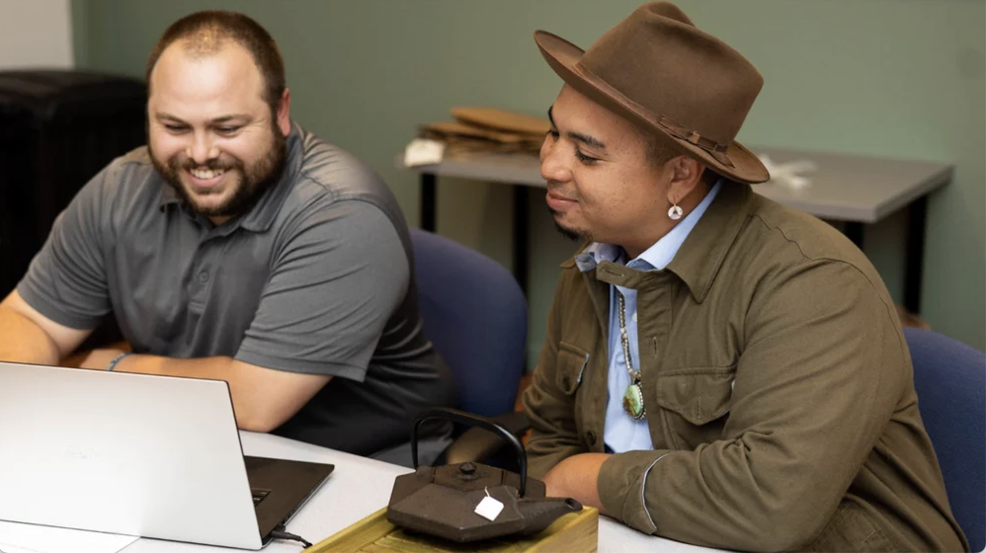 Two men sit smiling looking at a laptop screen during a digital coaching session focused on Indigenous-owned businesses. One wears a dark gray collared t-shirt and the other is wearing a brown brimmed hat and jacket, and a wampum earring and turquoise medallion with silver cord seeds representing his tribe and the Southwest.