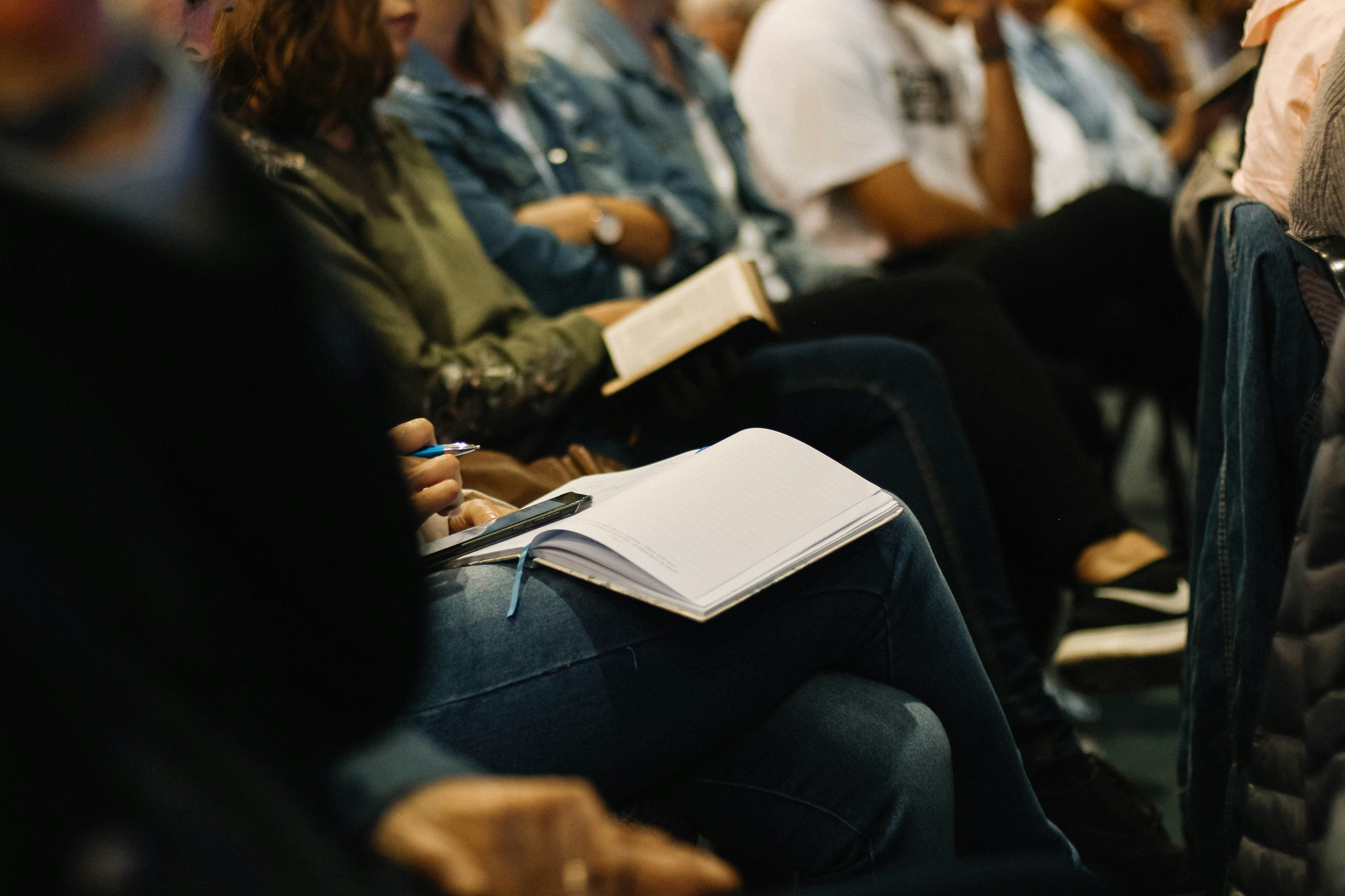 Row of people sitting with notebooks sitting open on the knees of the first two people visible 