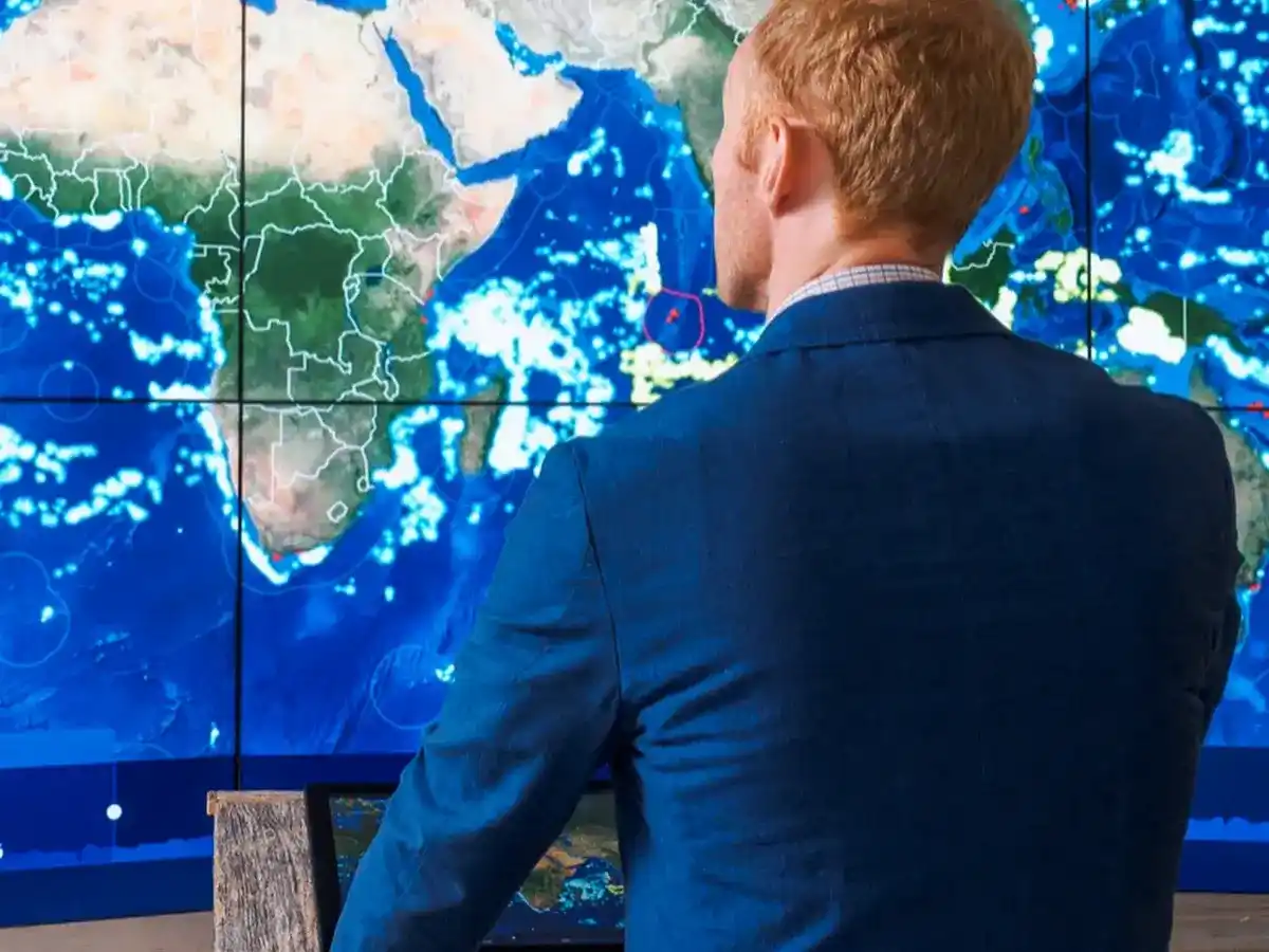 A man in a blue suit stands in front of a large digital map of the Earth.