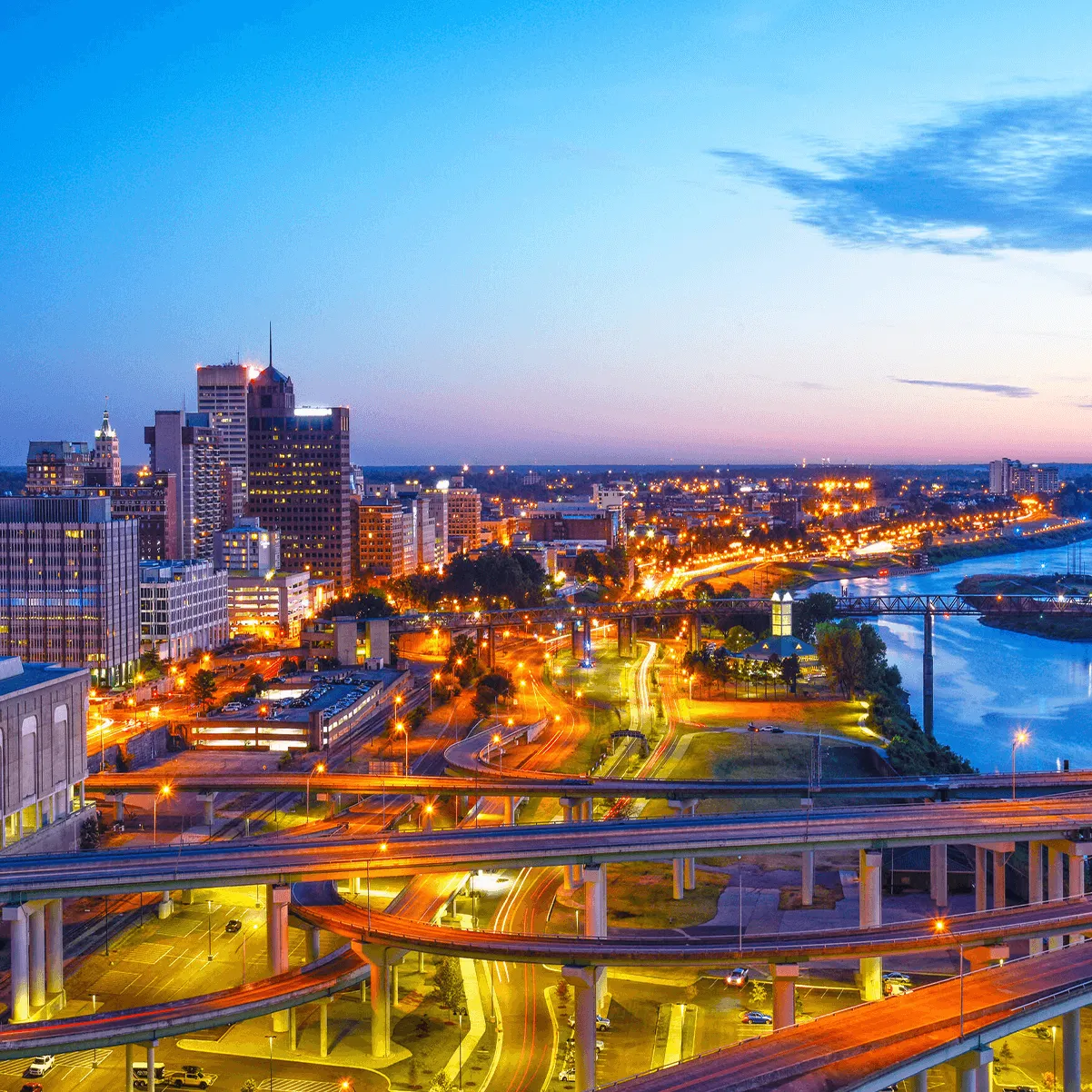 A cityscape of Memphis, Tennessee at dusk, showcasing illuminated buildings, highways, and the Mississippi River.