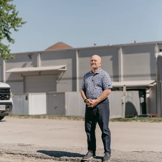 The president of a non-profit that feeds families in need, stands outside an industrial building with his hands clasped and a smile on his face.