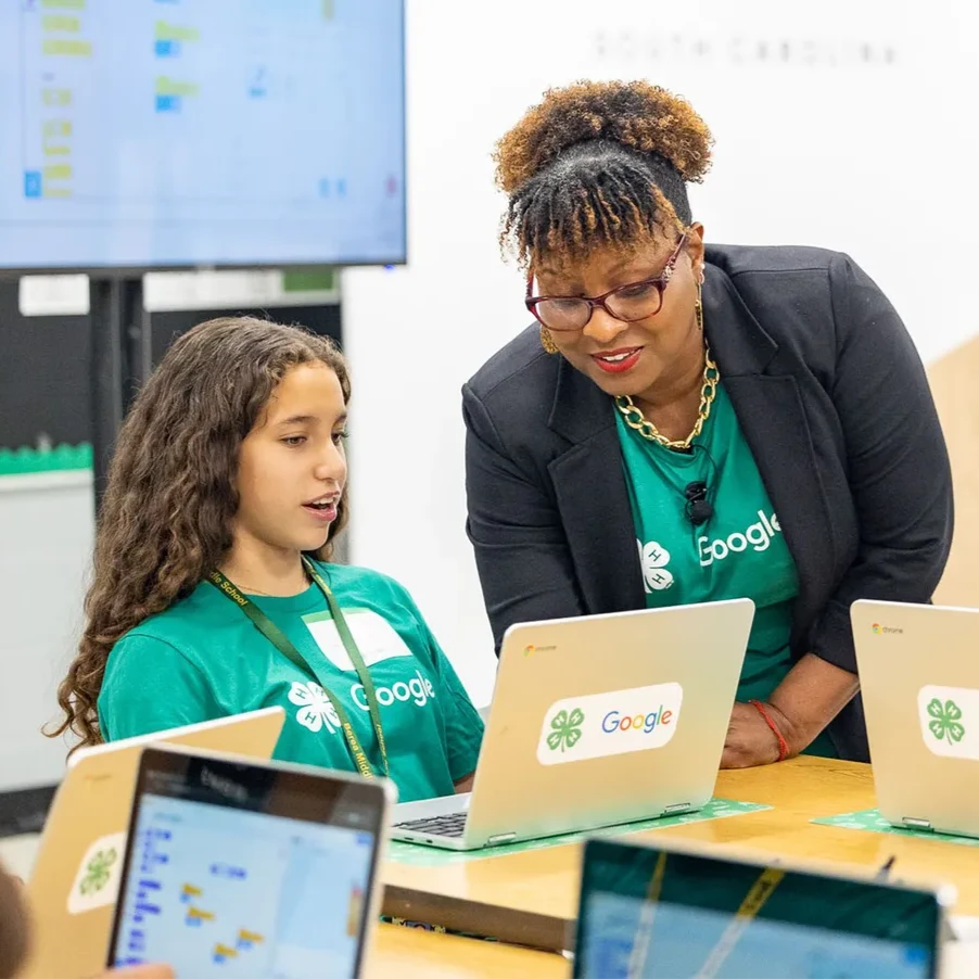 A teacher wearing a green shirt is leaning in to show a student something on a laptop