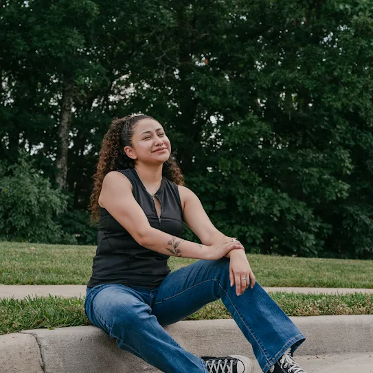 A recent Google Skilled Trades and Readiness Program graduate sits outside on the curb, smiling.