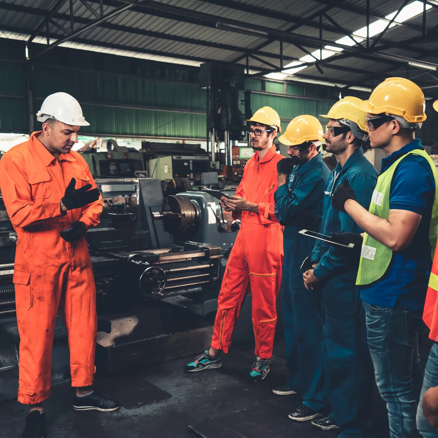 A group of workers standing around discussing something in the factory, all wearing hard hats and safety wear.