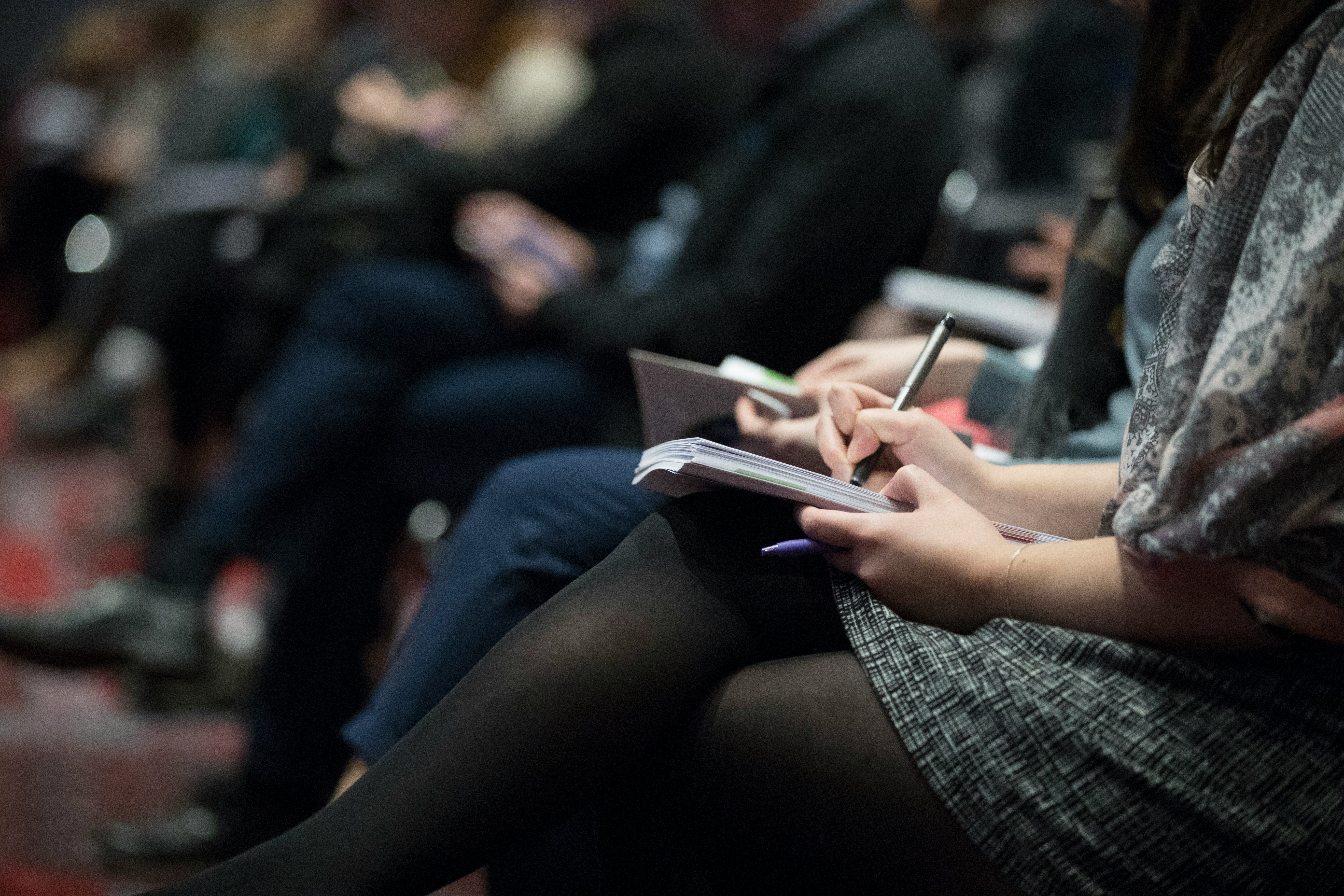 Row of people seated, with the first two people visible taking notes with pen and notepad