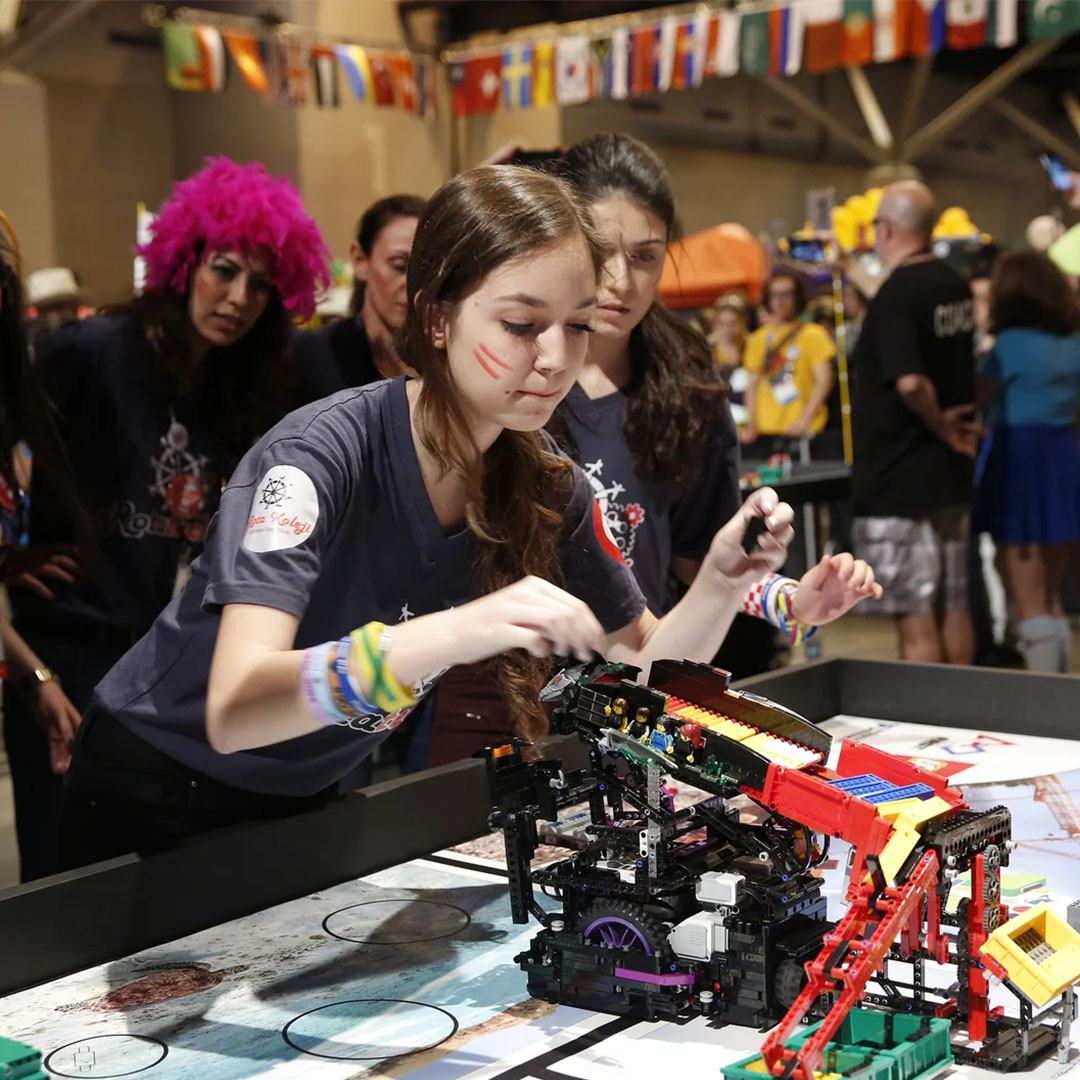 Young woman being cheered on by her teammates while she works on a small robot made of Legos