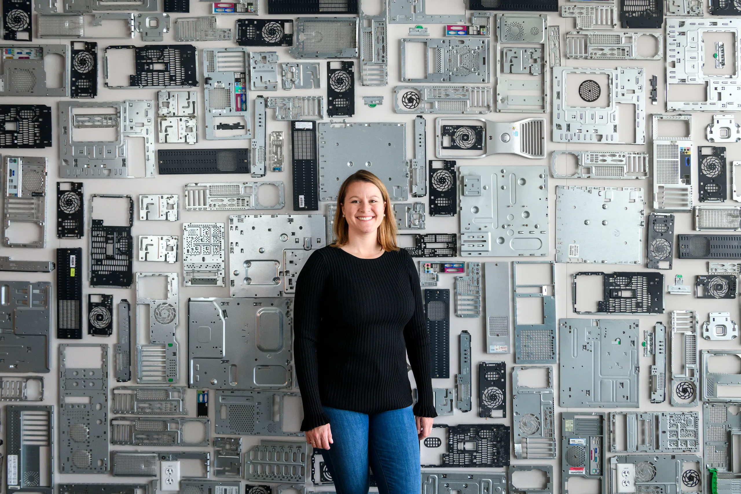 Photograph of a woman smiling standing in front of a wall covered in computer hardware.