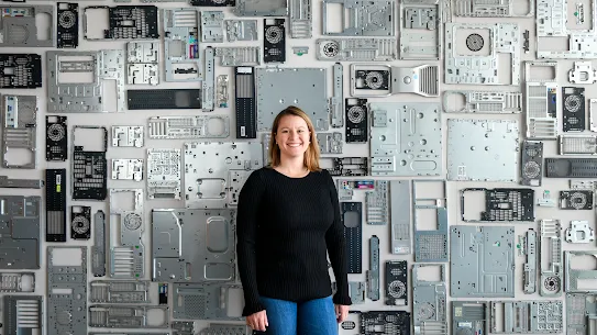 Photograph of a woman smiling standing in front of a wall covered in computer hardware.