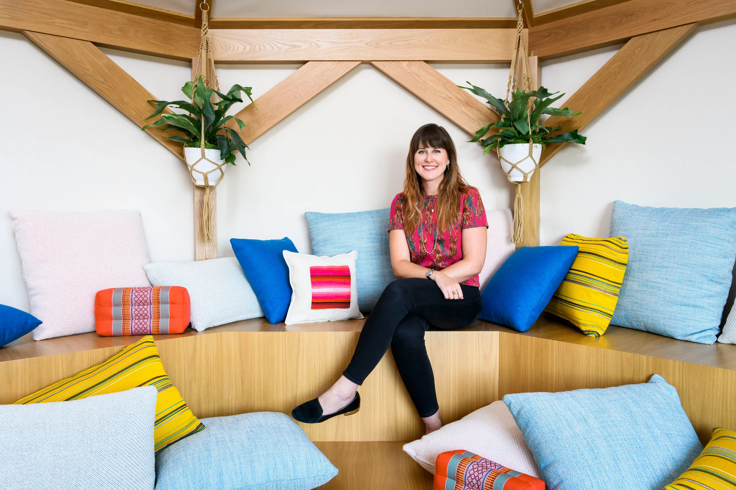 Photograph of a woman smiling sitting in a room filled with pillows and plants.