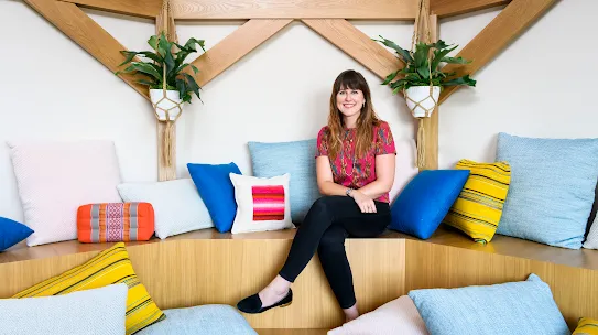 Photograph of a woman smiling sitting in a room filled with pillows and plants.