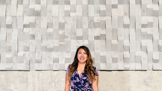 Photograph of a woman smiling standing in front of a textured wall.