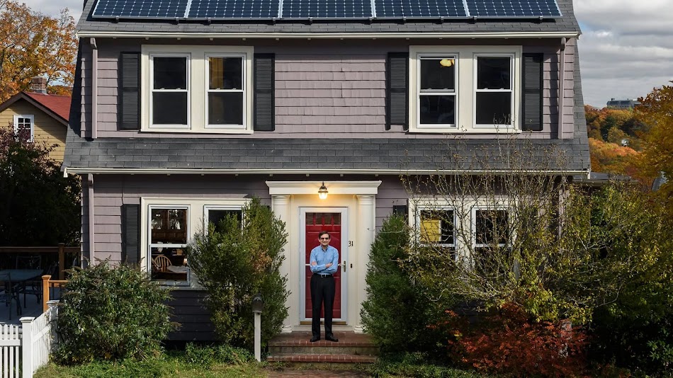 A man stands in front of a quaint home with solar panels on the roof.