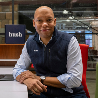  A man looking directly at the camera wearing a blue vest with the word ‘Hush’ on the left side, over a blue button down shirt, in an office setting