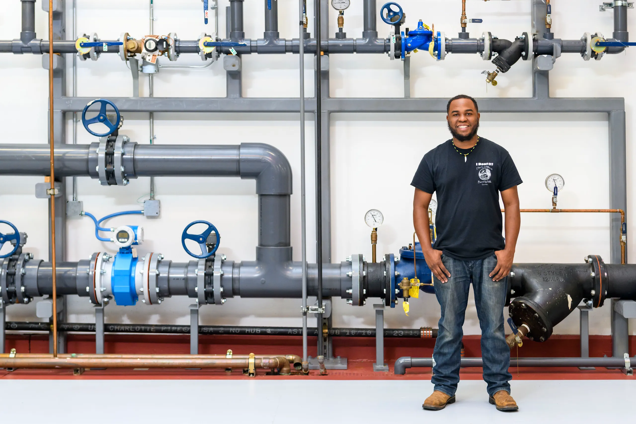 The image is a full-body photograph of a man in a black tshirt standing in front of pipes at a Google office.