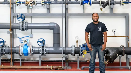 The image is a full-body photograph of a man in a black tshirt standing in front of pipes at a Google office.