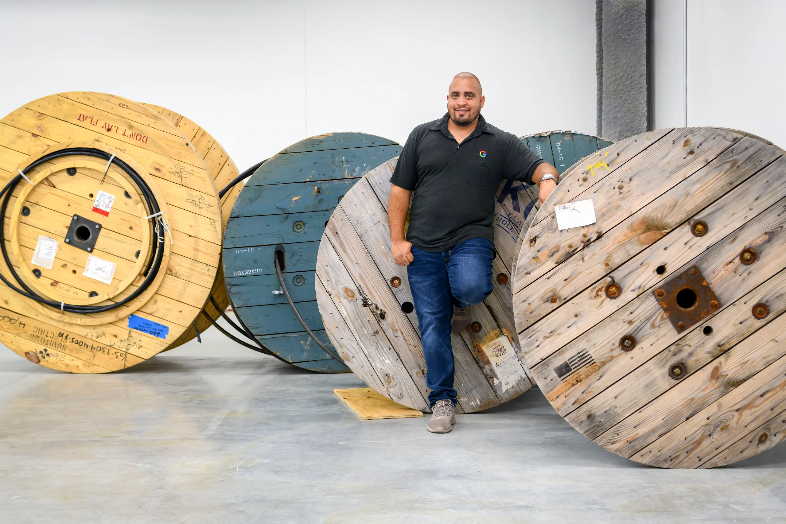 A full body photograph of a man in a black Google branded golf shirt standing amidst large wood wheels containing cables. 