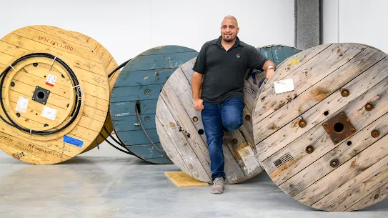 A full body photograph of a man in a black Google branded golf shirt standing amidst large wood wheels containing cables. 