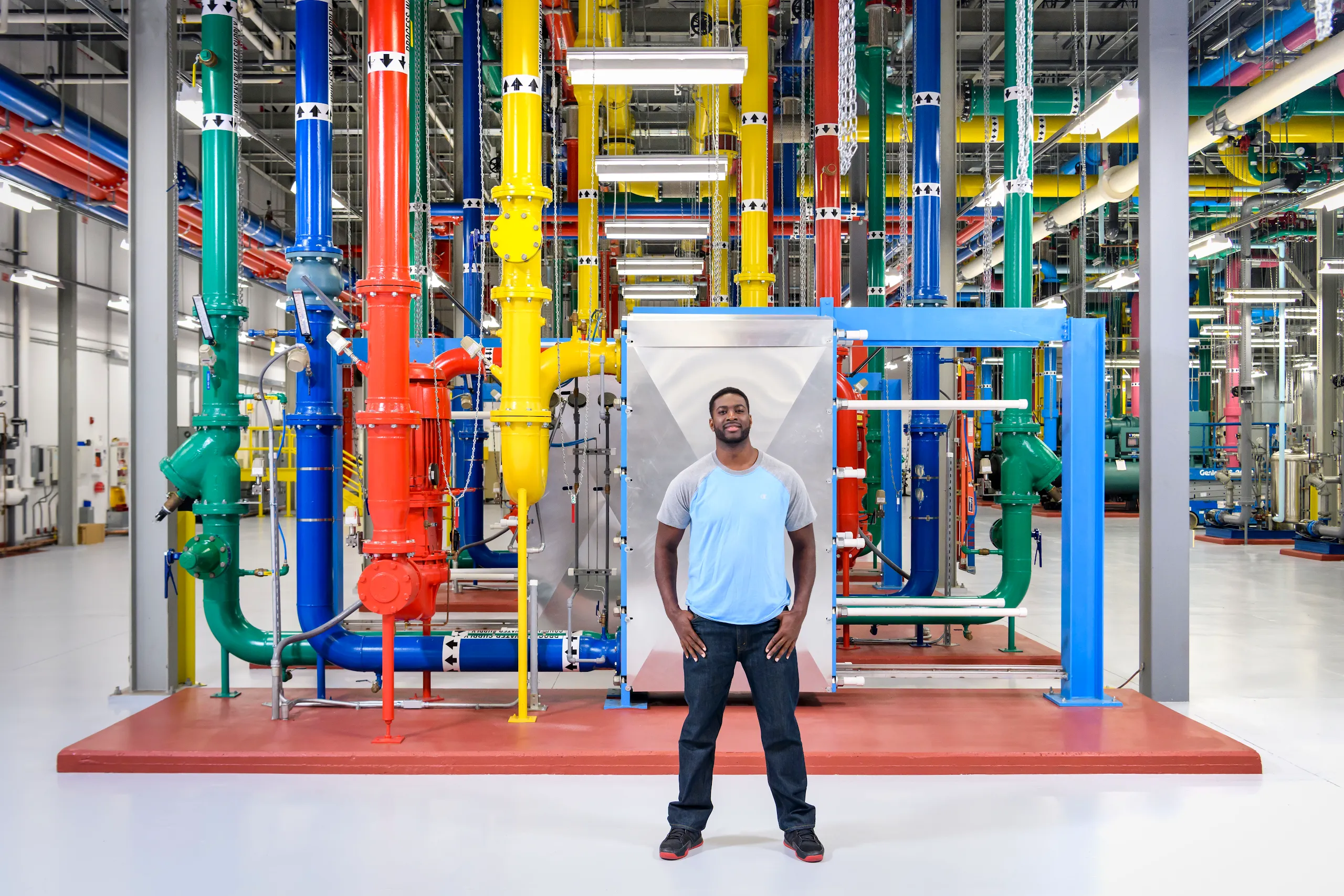 A photograph of a man with in a blue shirt with hands in pocket standing in the center of the image in front of colorful pipes in a Google facility. 