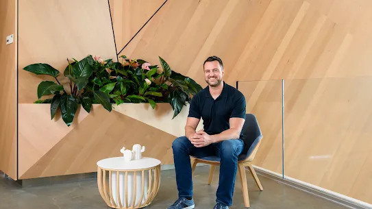 Photograph of a smiling man sitting in a chair in a room with wood paneling. 