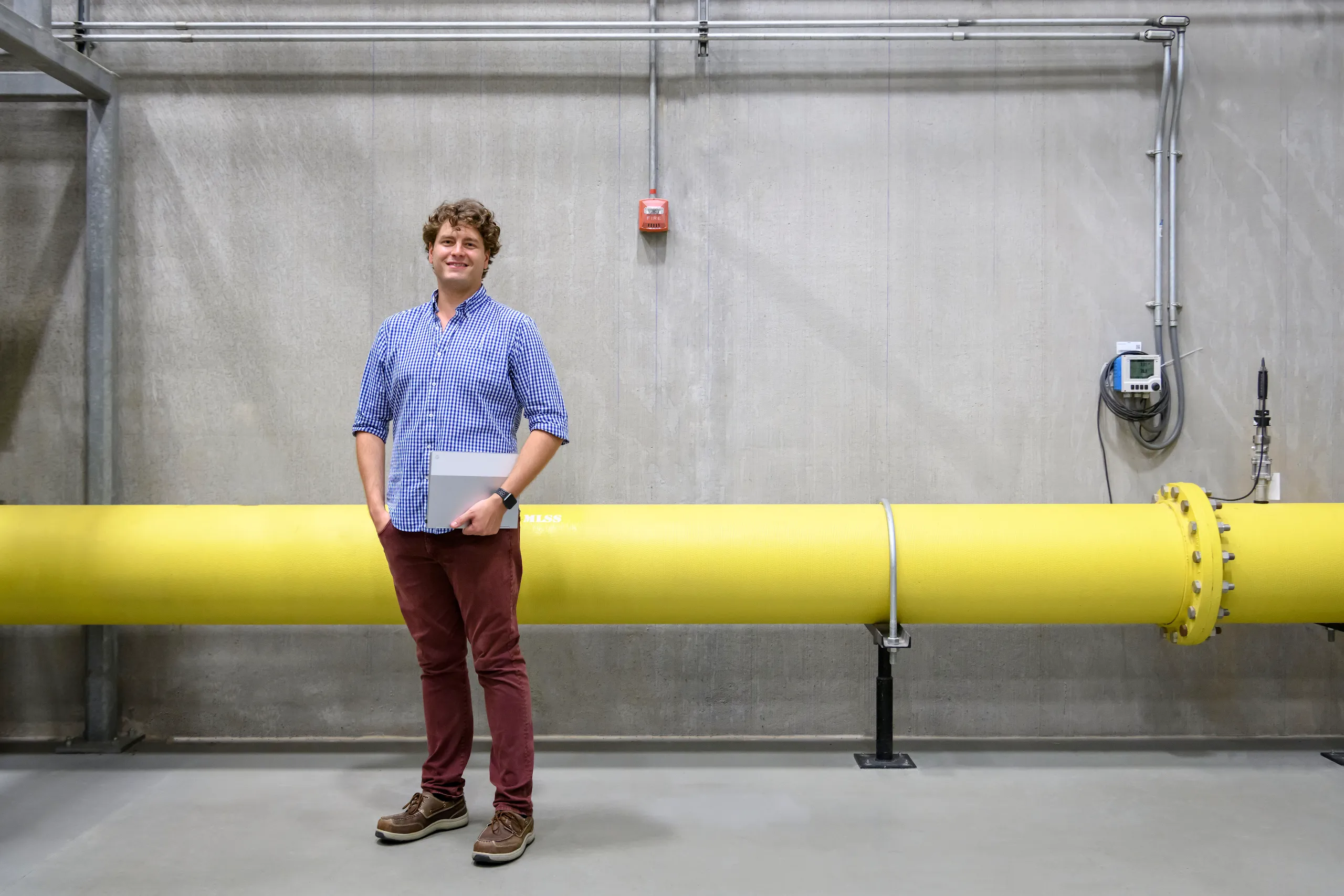 The image is a full-body photograph of a man in a blue and white checked shirt holding a folder in front of a yellow pipe at a Google office.