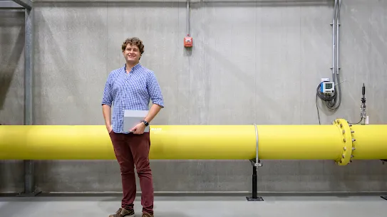 The image is a full-body photograph of a man in a blue and white checked shirt holding a folder in front of a yellow pipe at a Google office.