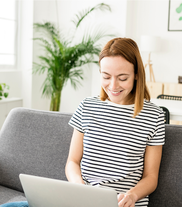 A young woman in a black and white striped top sits on a gray couch with an open laptop in her lap. She smiles as she types.