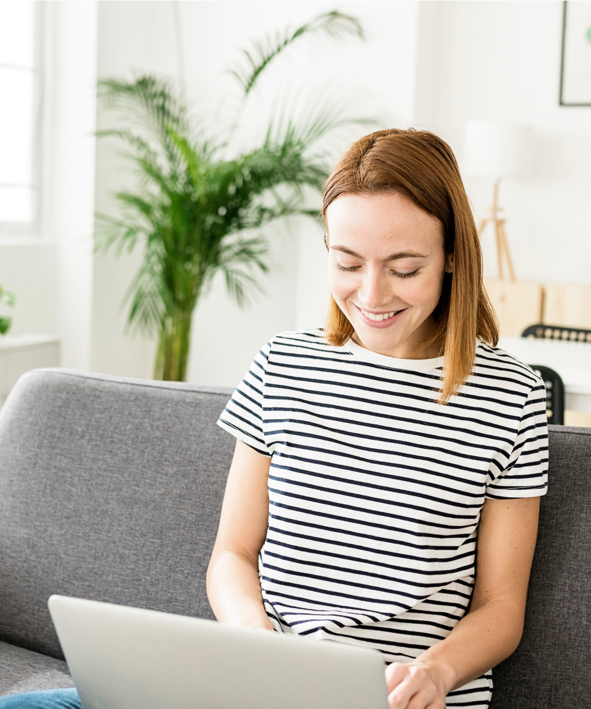 A young woman in a black and white striped top sits on a gray couch with an open laptop in her lap. She smiles as she types.